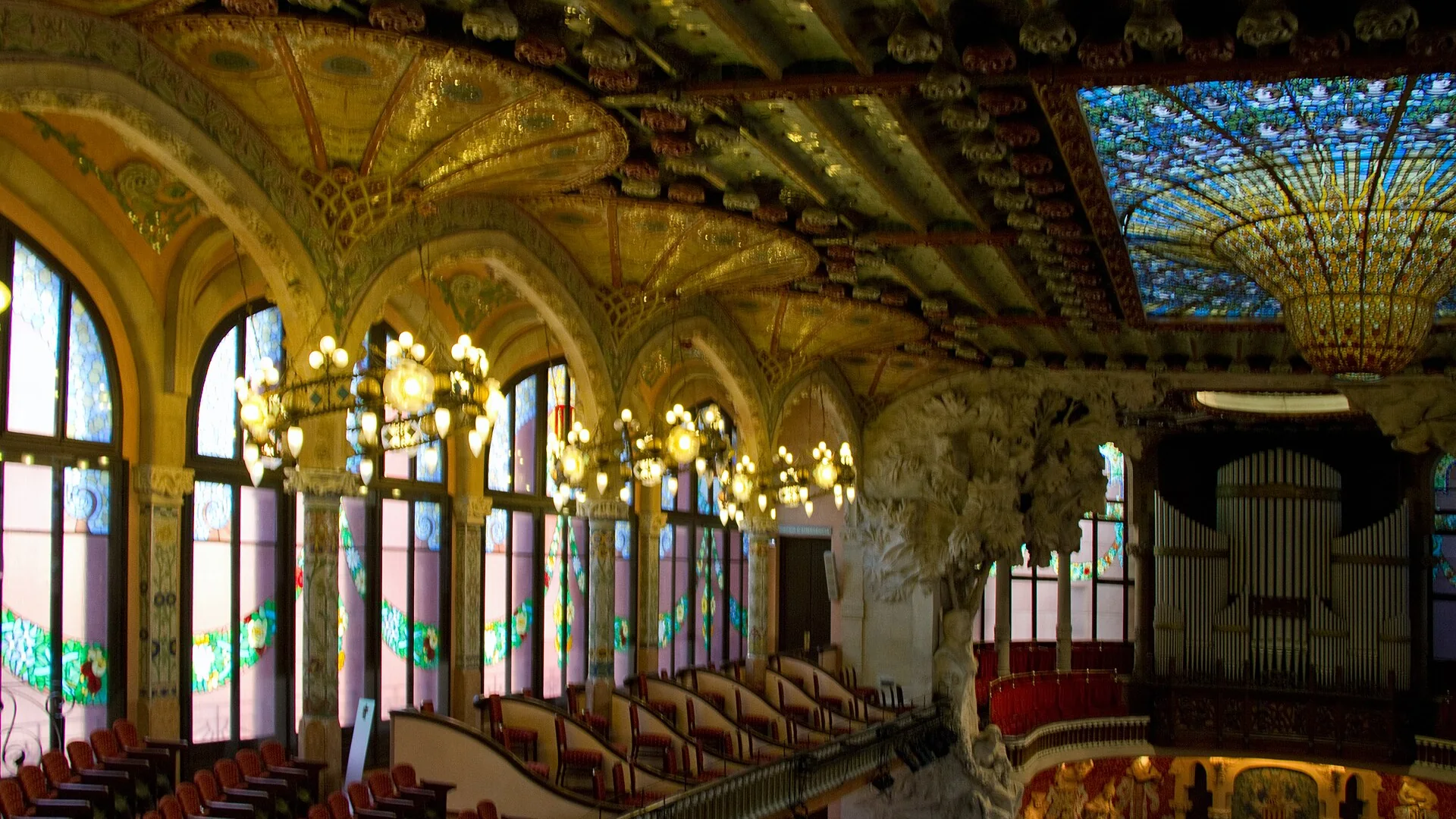 The concert hall of the Palau de la Música Catalana seen from the side: arched stained-glass windows, hanging lamps, a stained-glass skylight in gold and blue, and the stage with its sculpted muses and organ.