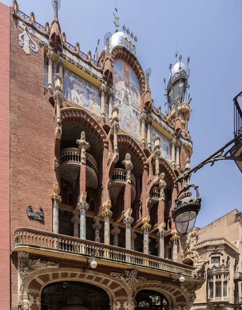 The corner front of the Palau de la Música Catalana in red brick, with mosaic columns, balconies and a sculpted pediment against a blue sky.