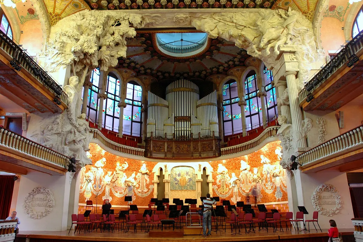 The stage of the Palau concert hall with white sculpted trees, the organ above and a mosaic wall of muses behind the chairs of an orchestra.